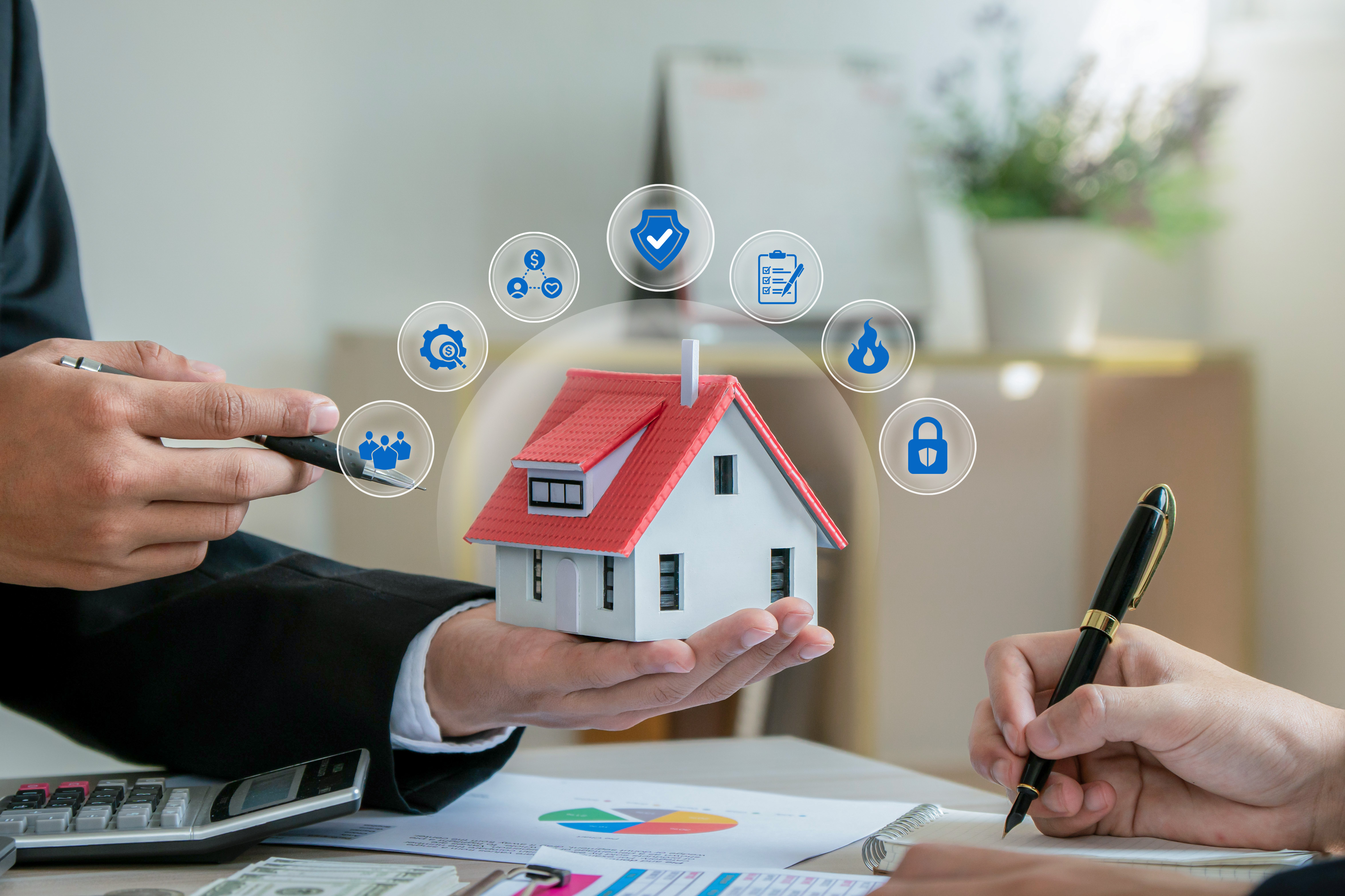 a person holding a small model house with floating icons around it. While another person is writing, with business documents, a calculator, and cash on the table.